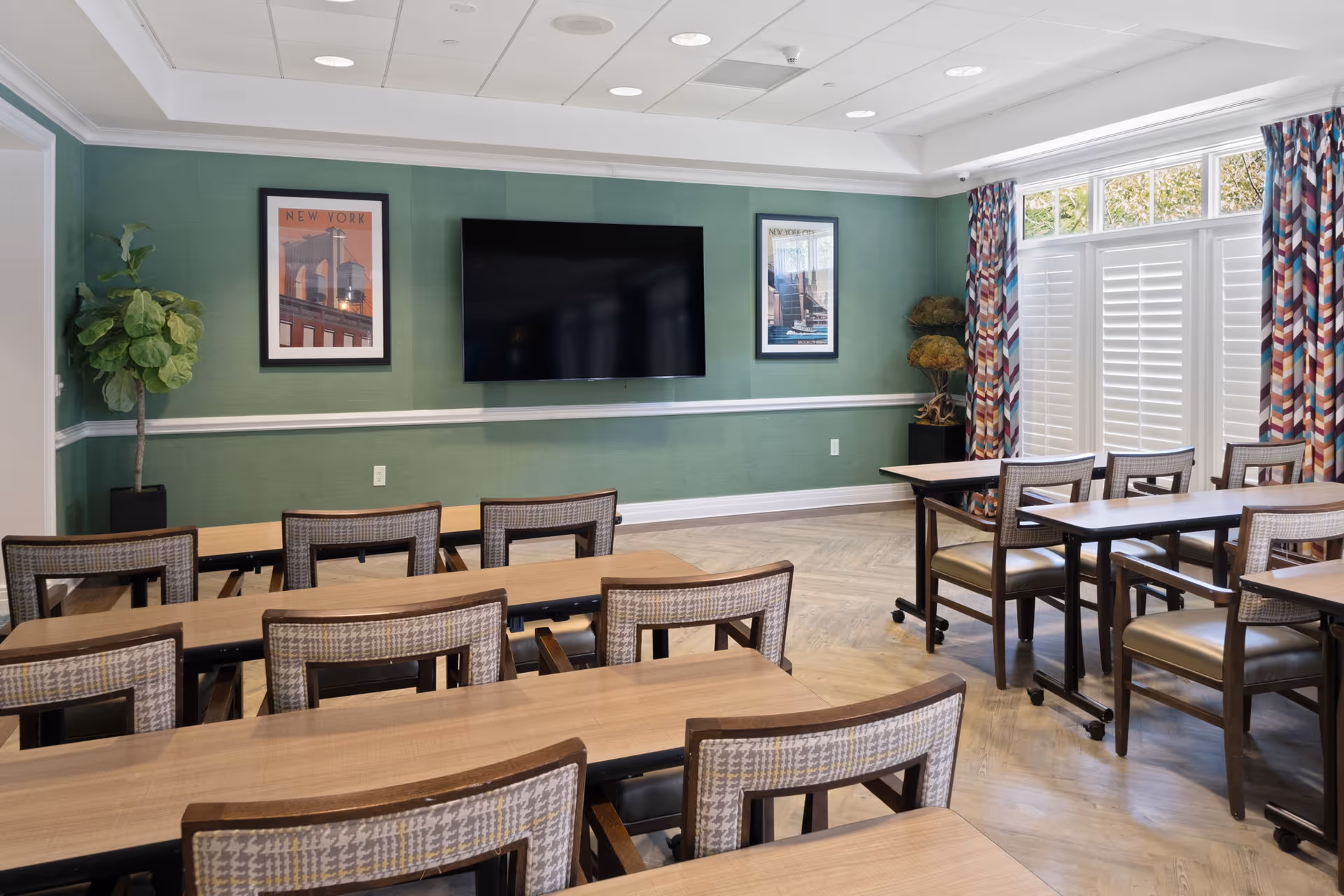 A well-lit room with multiple wooden tables and chairs arranged in rows facing a large flat-screen TV mounted on a green wall. The room has two framed pictures on the wall, patterned curtains on the windows, and two potted plants in the corners.