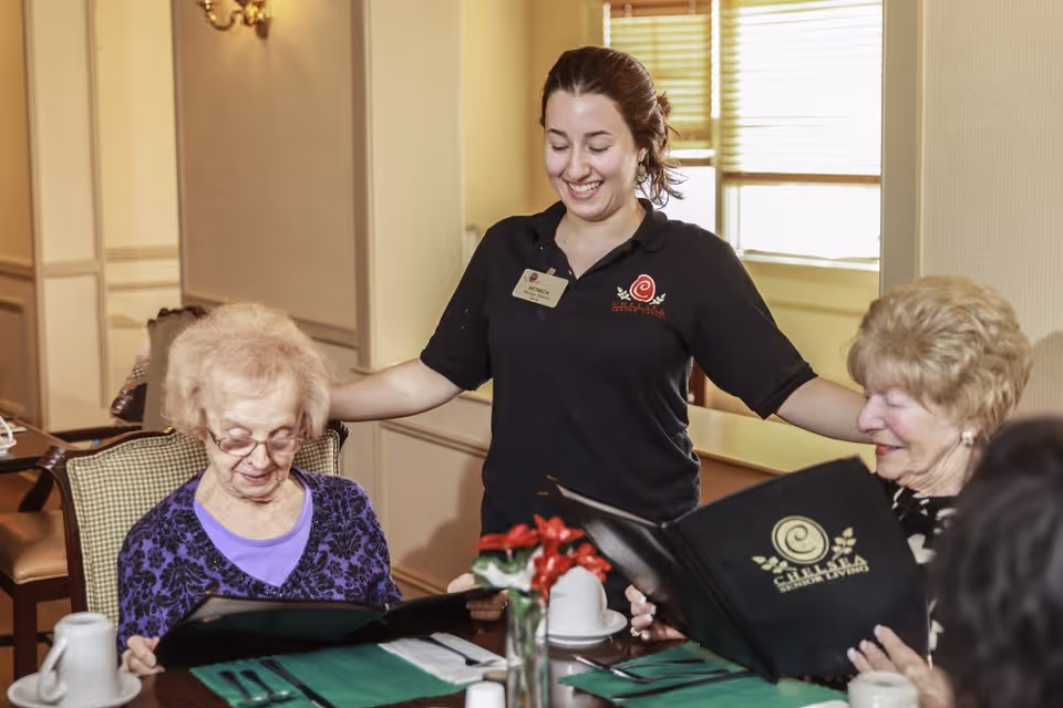 A smiling staff member in a black polo shirt with a name tag stands behind two elderly women seated at a dining table, looking at menus in a well-lit room with beige walls and a window with blinds.