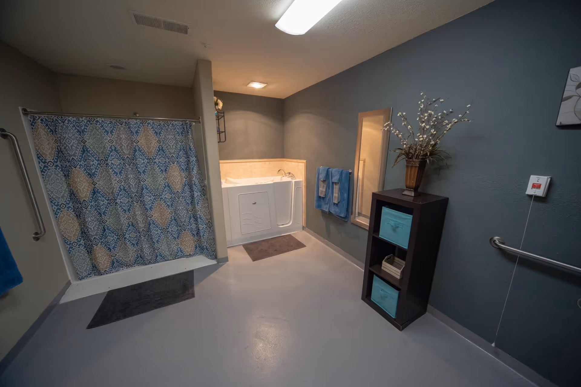 A spacious bathroom with a walk-in bathtub, a shower area with a patterned curtain, blue and beige towels hanging on the wall, a tall black shelving unit with blue storage bins and a vase with flowers, and grab bars on the walls for safety.