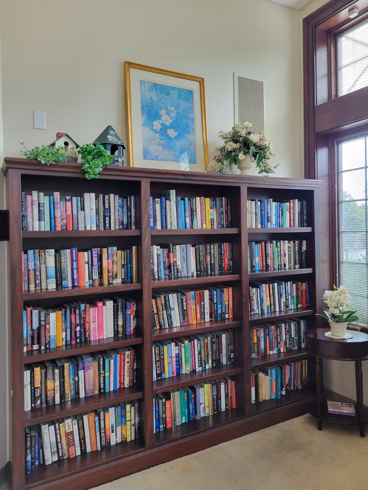 A wooden bookshelf filled with numerous books, positioned against a light-colored wall. On top of the bookshelf are two decorative birdhouses, a green leafy plant, and a vase with white and pink flowers. Above the bookshelf hangs a framed painting with a gold frame depicting blue and white flowers. To the right of the bookshelf is a window with wooden trim and blinds, and a small round wooden table with a white flower arrangement on it.