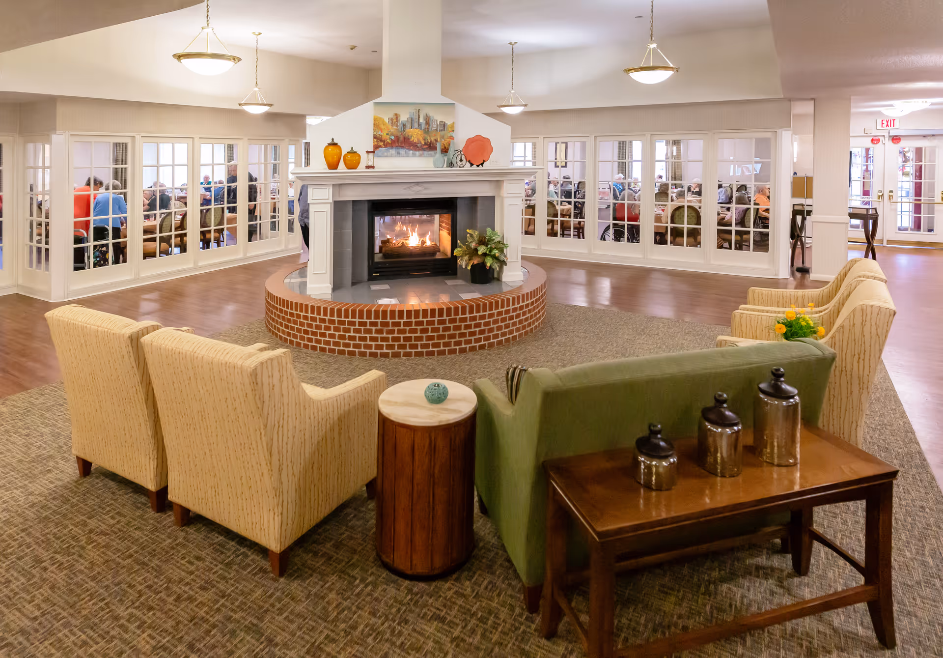 A cozy senior living facility common area with a central brick fireplace surrounded by comfortable armchairs and a green sofa. Behind the fireplace, a dining room with large windows shows several seniors seated at tables. The room is well-lit with ceiling lights and decorated with vases and a painting above the fireplace.