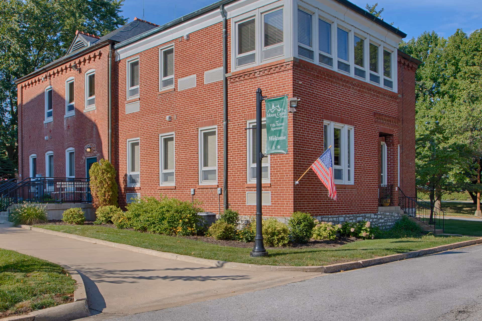 Red brick two-story senior facility building with an American flag and a welcome sign on a lamp post, surrounded by landscaping.