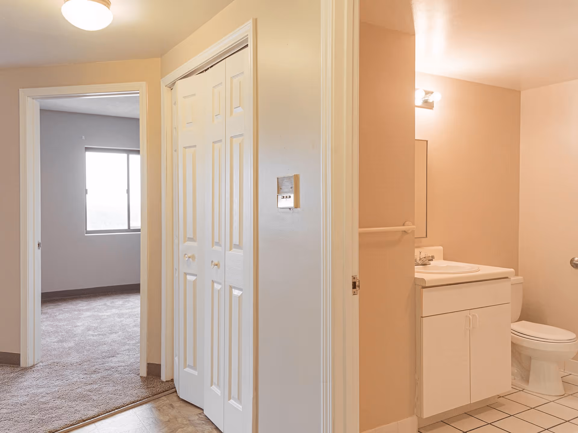 Interior view of a hallway in a senior living facility showing a bathroom on the right with a white vanity, sink, mirror, and toilet. On the left, there is an open doorway leading to a carpeted bedroom with a window. The hallway has a closet with white bi-fold doors.