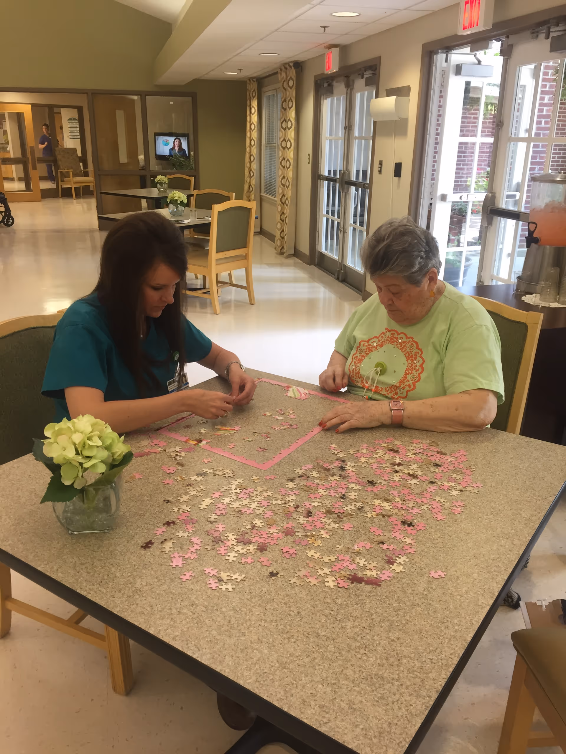 Two women sitting at a table working on a jigsaw puzzle together in a well-lit common area with chairs, tables, and large windows. One woman is wearing a green shirt and the other is in a teal uniform. There is a small vase with flowers on the table.