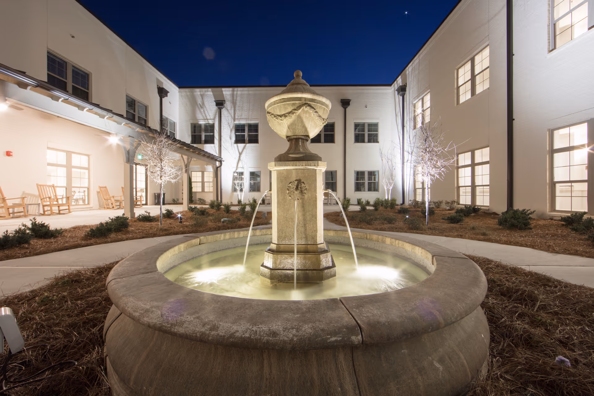 Night view of a courtyard at The Blake at Woodcreek Farms featuring a large stone fountain with water flowing from four spouts into a circular basin. The courtyard is surrounded by a two-story white building with multiple windows and a covered patio area with rocking chairs. Small trees and shrubs are planted around the fountain, and the area is softly illuminated by outdoor lighting.