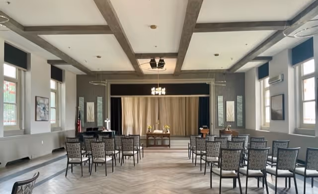 Bright multipurpose meeting room with rows of chairs facing a small stage and podium beneath exposed ceiling beams.