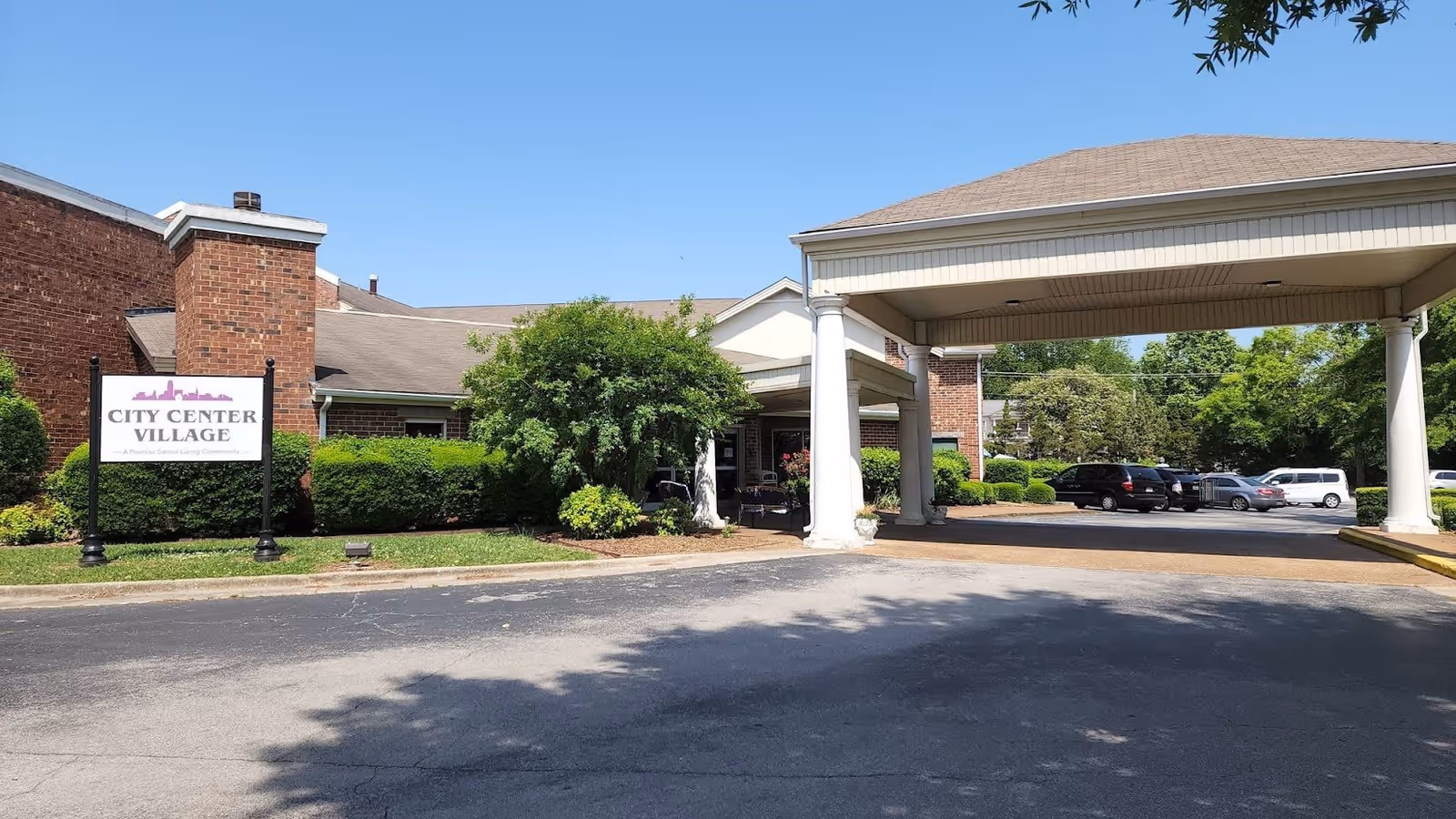 Exterior view of City Center Village Senior Community showing the entrance with a covered drop-off area supported by white columns, a parking lot with several cars, and a sign with the facility's name on a lawn with bushes and trees under a clear blue sky.