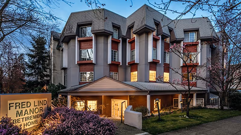 Exterior view of Fred Lind Manor retirement home building during dusk, showing a multi-story structure with illuminated windows, surrounded by trees and landscaping, and a sign in front reading 'Fred Lind Manor Retirement Home'.