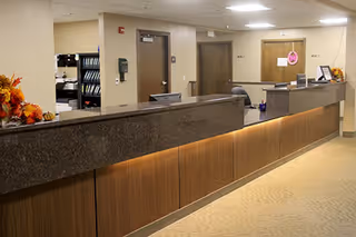 Reception desk area inside a senior living facility with a long granite countertop, wooden paneling below, and office doors in the background. The area is well-lit with ceiling lights and decorated with autumn-themed floral arrangements.