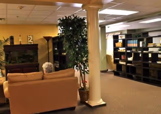 Interior view of a senior living facility common area featuring a beige sofa, a tall potted plant, a wooden cabinet with a fish tank, a floor lamp, and black shelving units filled with books and decorative items. The space has a beige carpet and a white ceiling with fluorescent lighting.