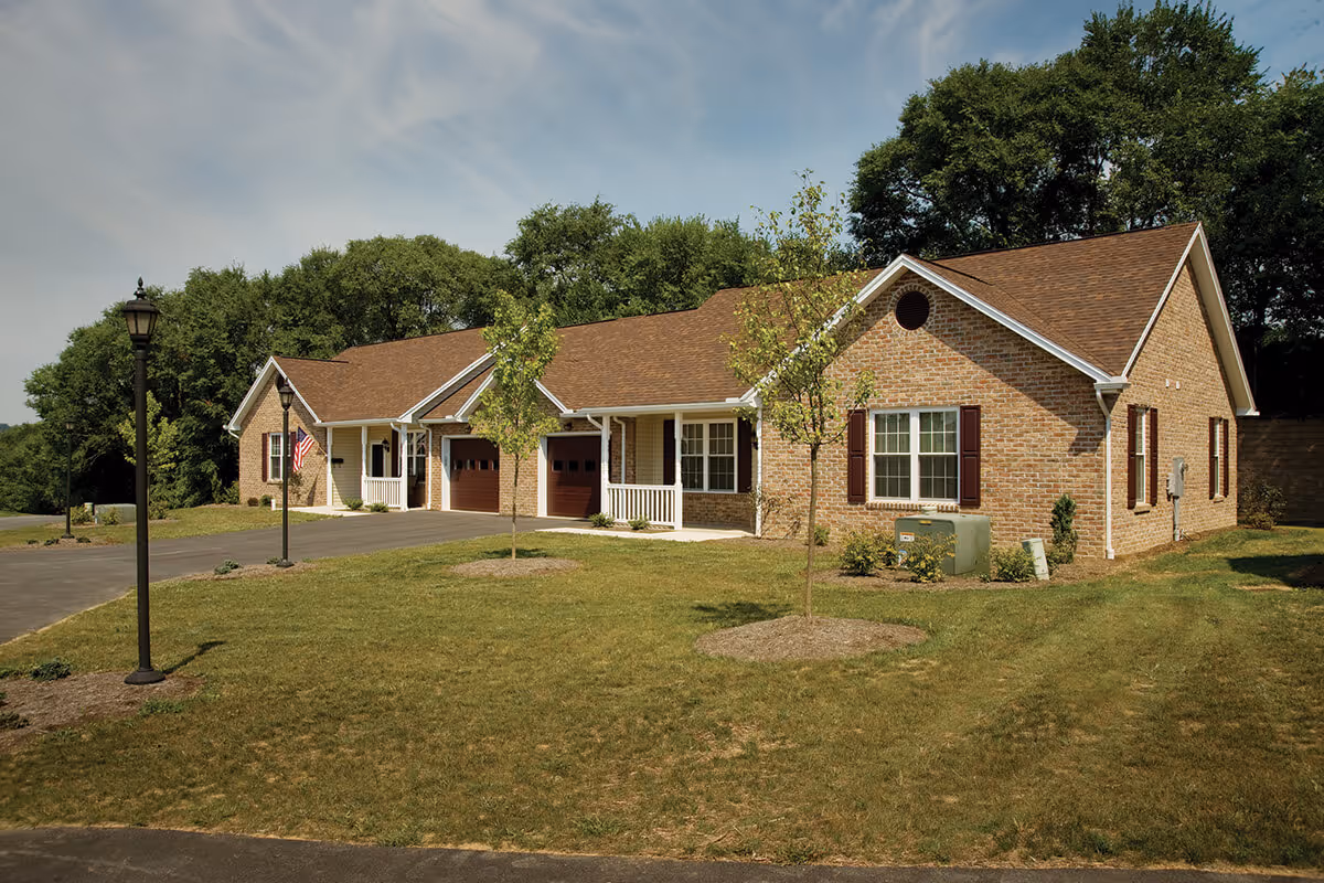 Single-story brick residential building with attached garages and porches on a grassy lawn with lamp posts and trees behind it.