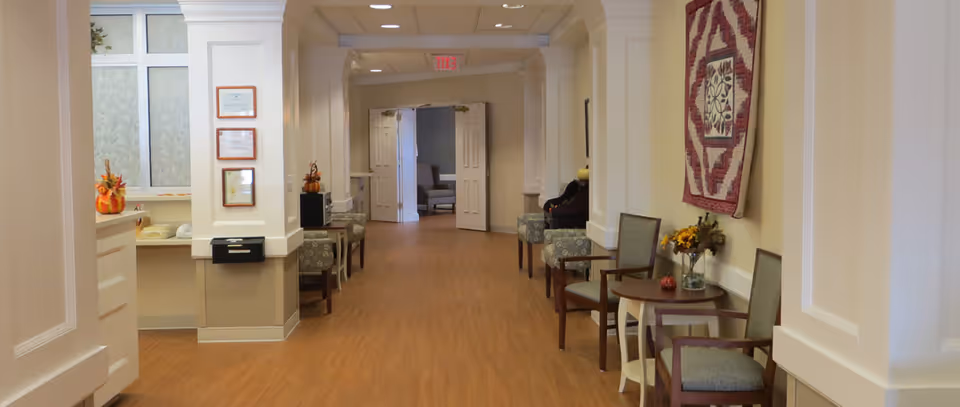 Wide interior hallway in a senior living community with chairs, small tables and wall decor leading to double doors.