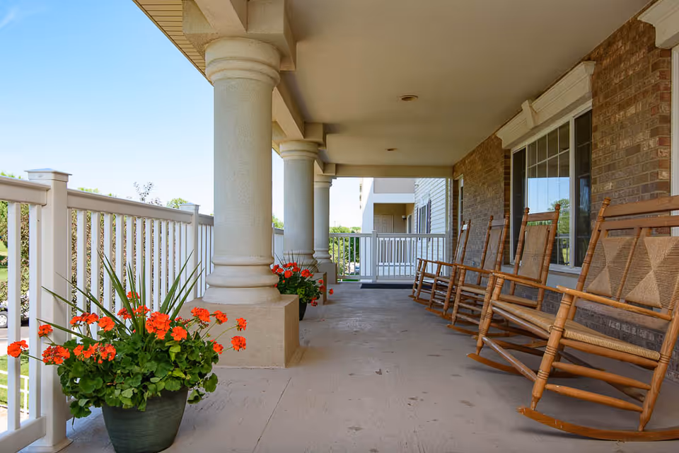 Covered front porch with wooden rocking chairs, columns, white railing and potted red flowers.
