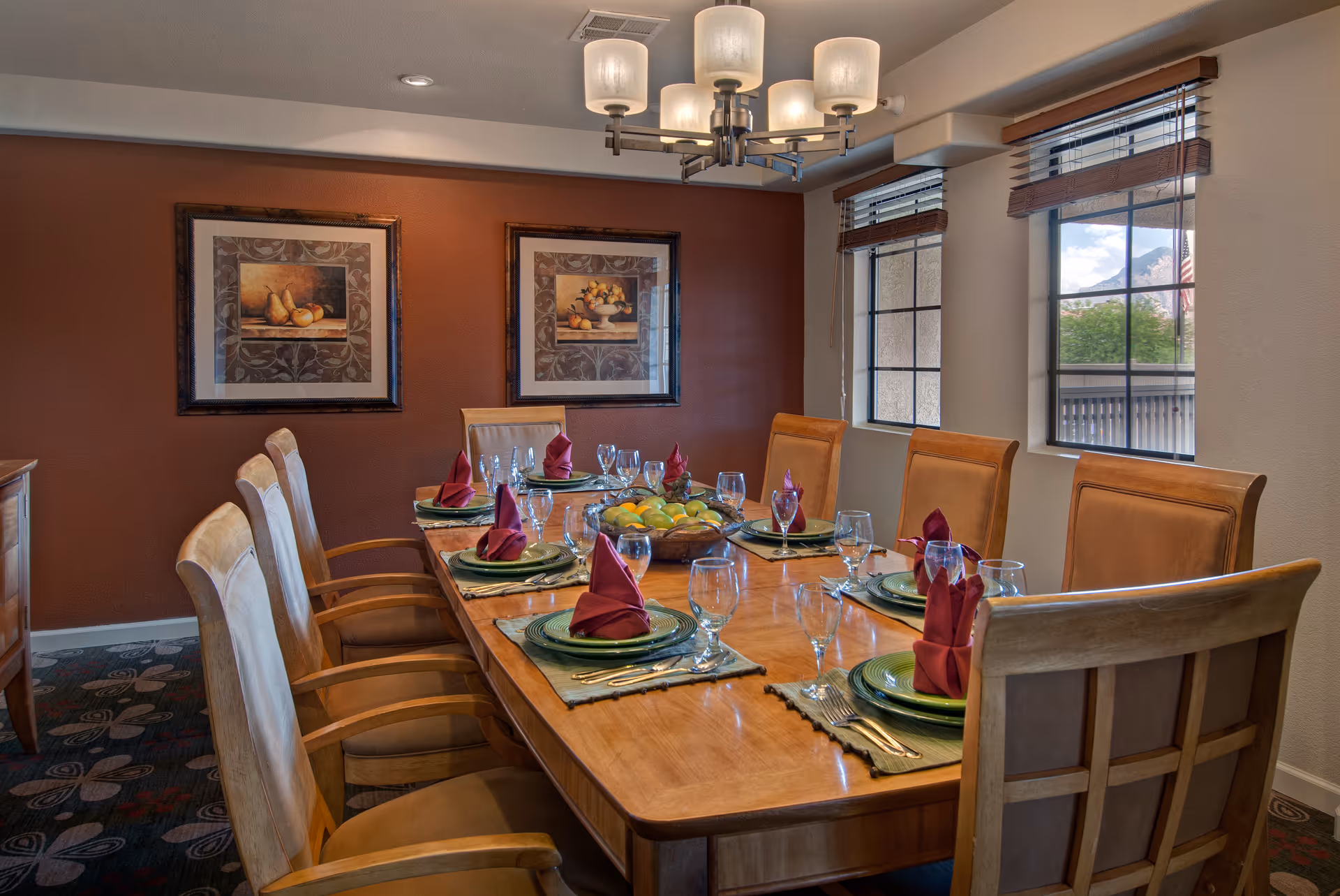 A dining room with a long wooden table set for eight people. Each place setting has a green plate, silverware, a wine glass, and a folded burgundy napkin. The room has two framed paintings of fruit on a dark red accent wall and two windows with wooden blinds letting in natural light. A modern chandelier with five lights hangs above the table.