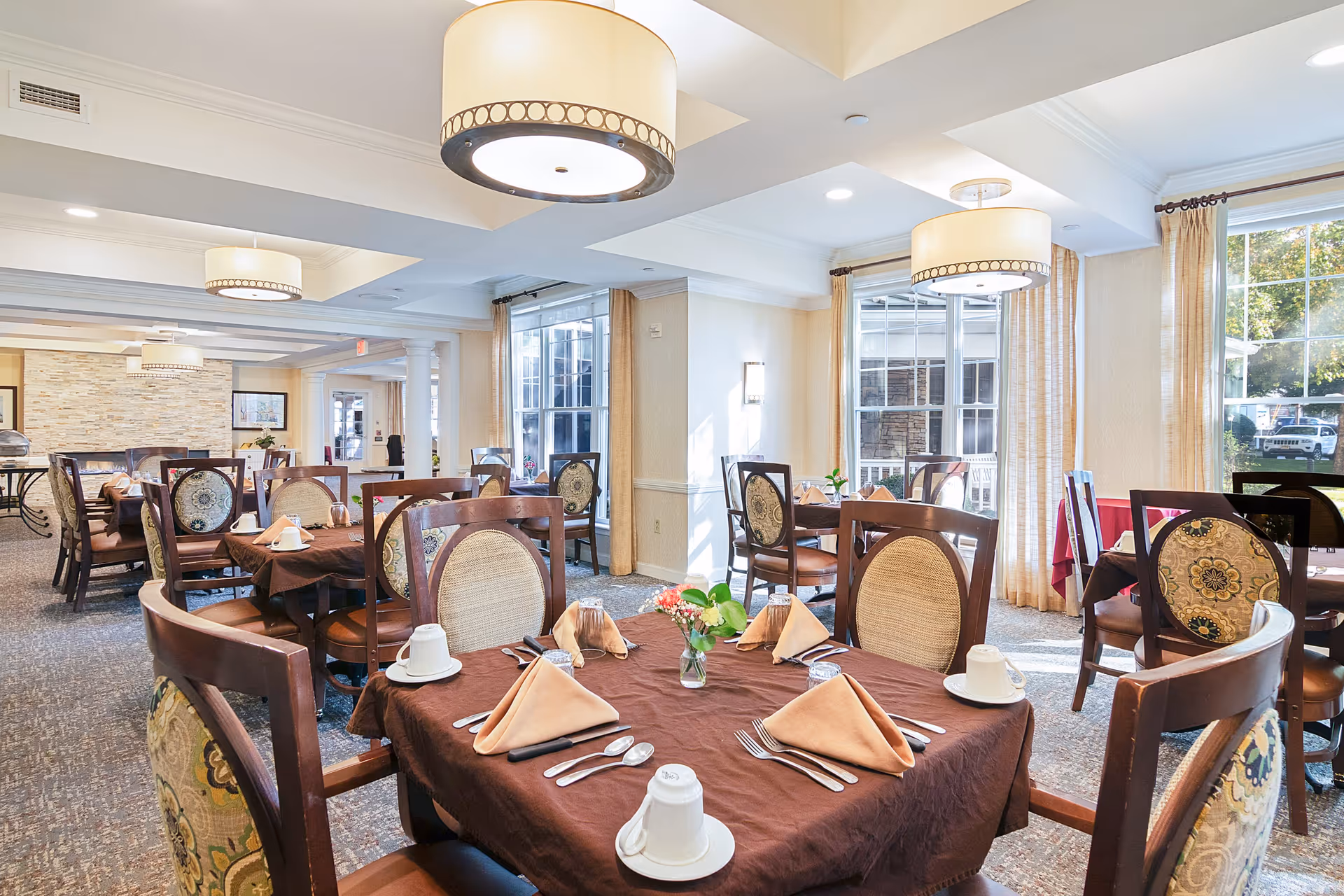 A bright and elegant dining room with several tables covered in brown tablecloths, each set with beige folded napkins, white cups, and silverware. The room features large windows with beige curtains allowing natural light to fill the space, patterned chairs, and modern ceiling lights.