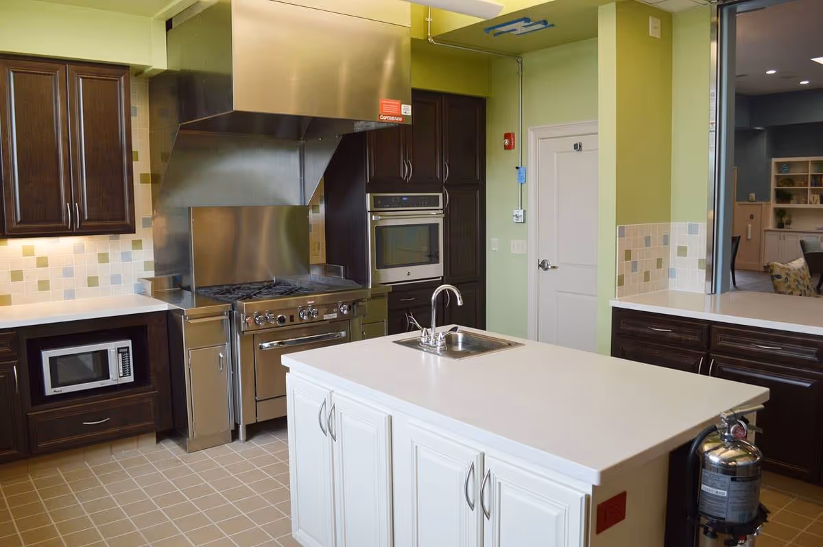 Modern kitchen with a large white island countertop featuring a built-in sink and faucet. The kitchen has dark wooden cabinets, a stainless steel stove with a large vent hood, a built-in microwave, and an oven. The walls are painted light green with a tiled backsplash in neutral tones. A fire extinguisher is mounted on the side of the island.