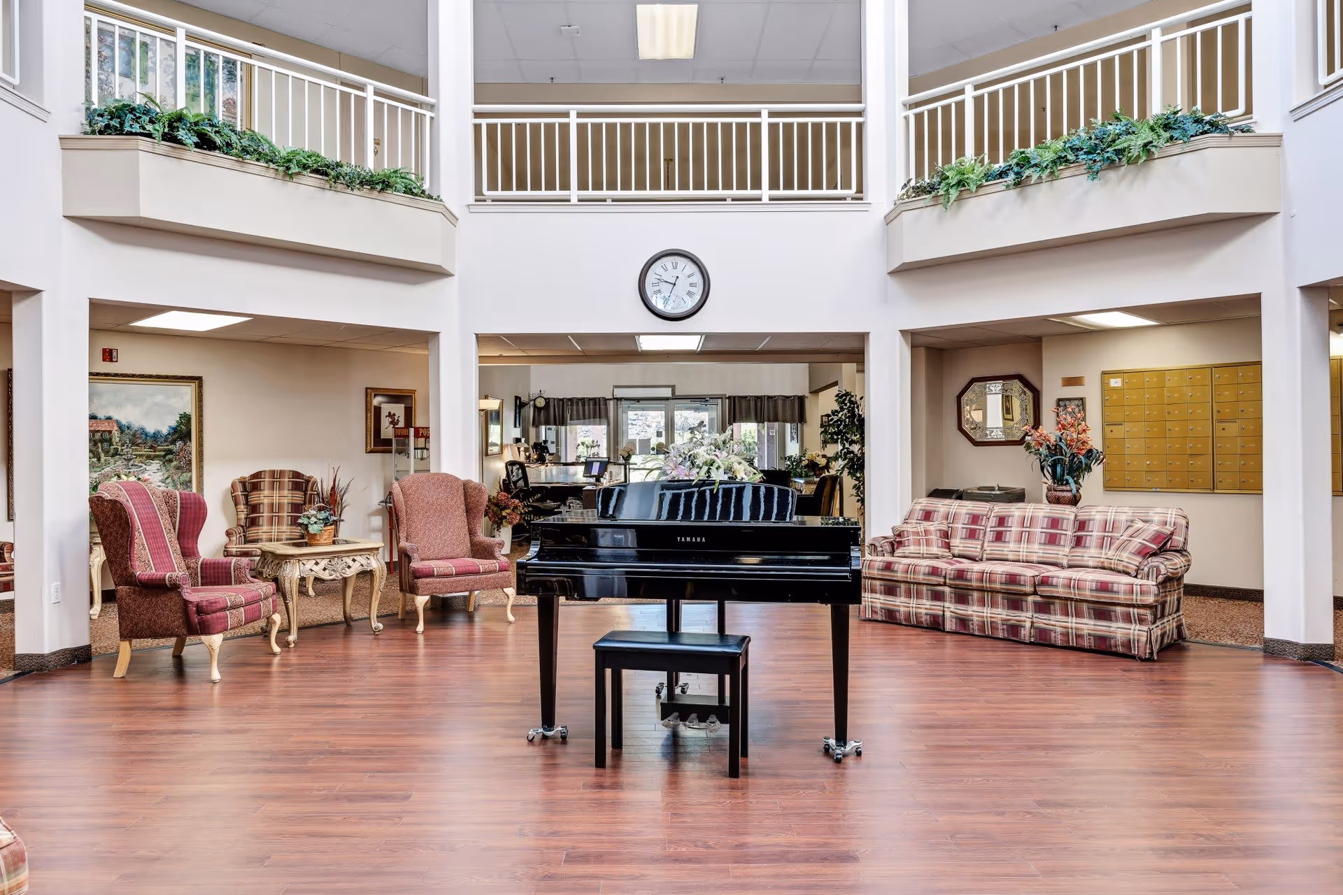 A spacious living room area with a polished wooden floor, featuring a black Yamaha grand piano with a matching bench in the center. Surrounding the piano are various seating options including a plaid sofa, two upholstered armchairs with red and beige patterns, and a small table with a plant. The room has white walls and a high ceiling with a second-floor balcony railing decorated with green plants. A clock is mounted on the wall above the piano, and there are decorative elements such as framed paintings and a mirror on the walls.