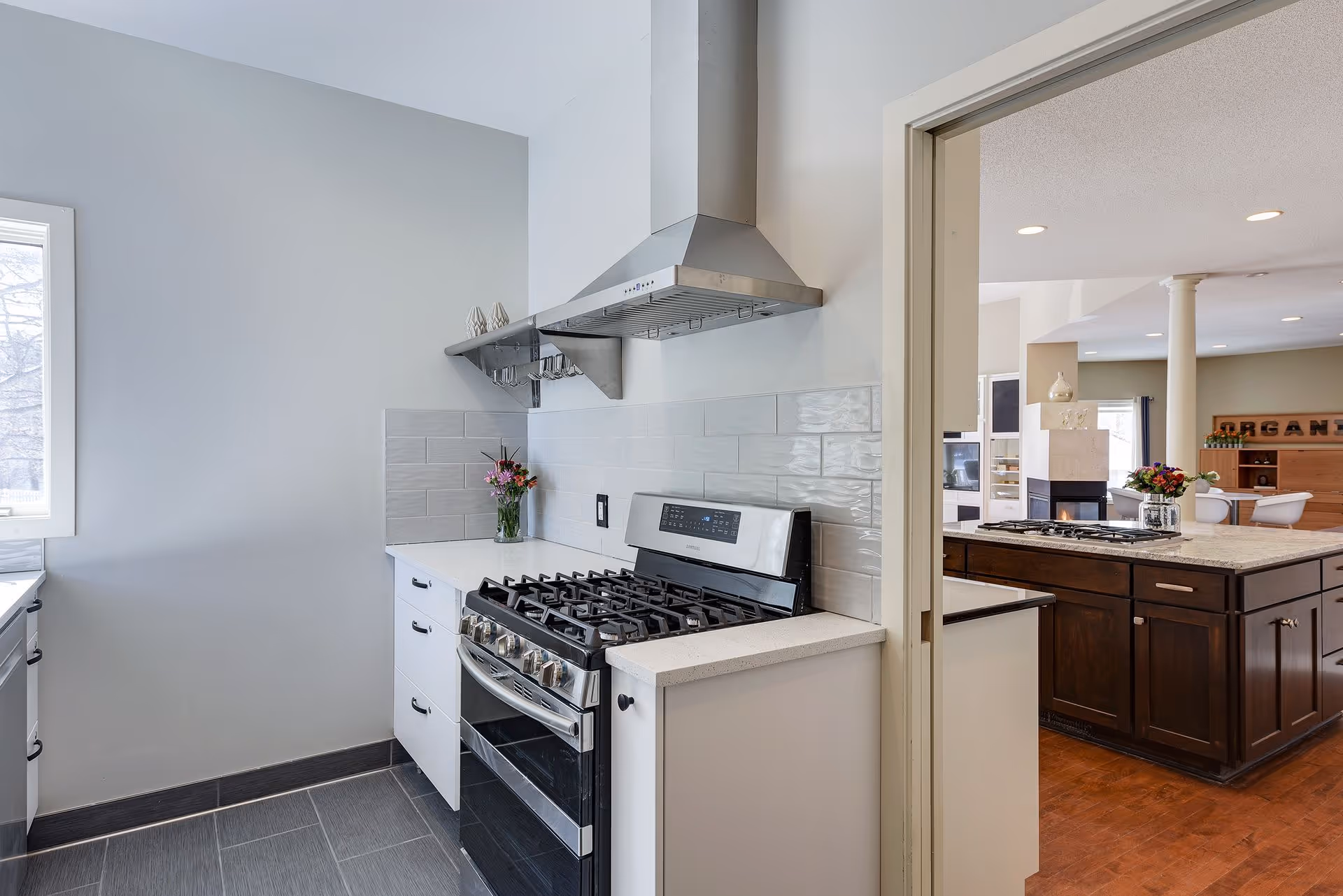 Modern kitchen area with a stainless steel gas stove and range hood, white cabinets, and a small vase with flowers on the counter. Through an open doorway, a larger kitchen island with a gas cooktop and dark wood cabinets is visible, along with a living area in the background.
