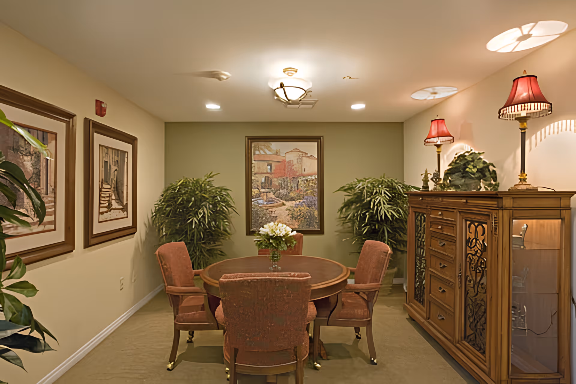 Small furnished dining room with a round wooden table surrounded by upholstered chairs, potted plants, framed artwork, and a wooden cabinet with lamps.