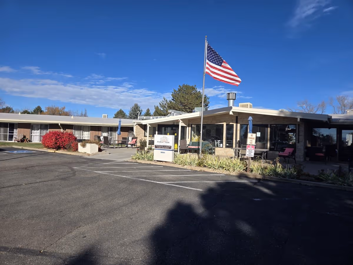 Front exterior of a single-story care facility with an American flag, entrance sign, outdoor seating, and a parking lot.