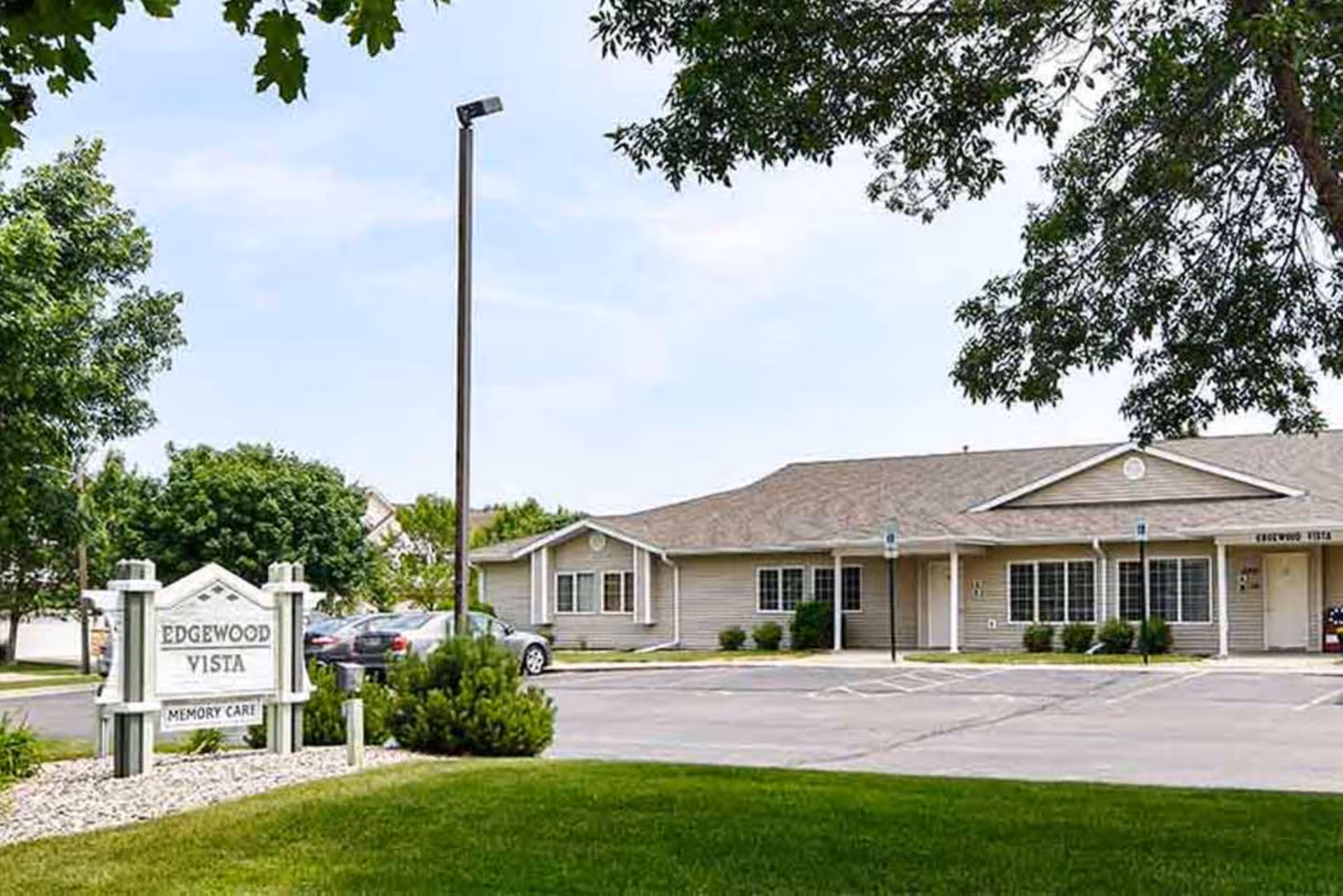 Single-story beige memory care building and parking lot with a white sign reading 'EDGEWOOD VISTA MEMORY CARE' on a manicured lawn and trees.
