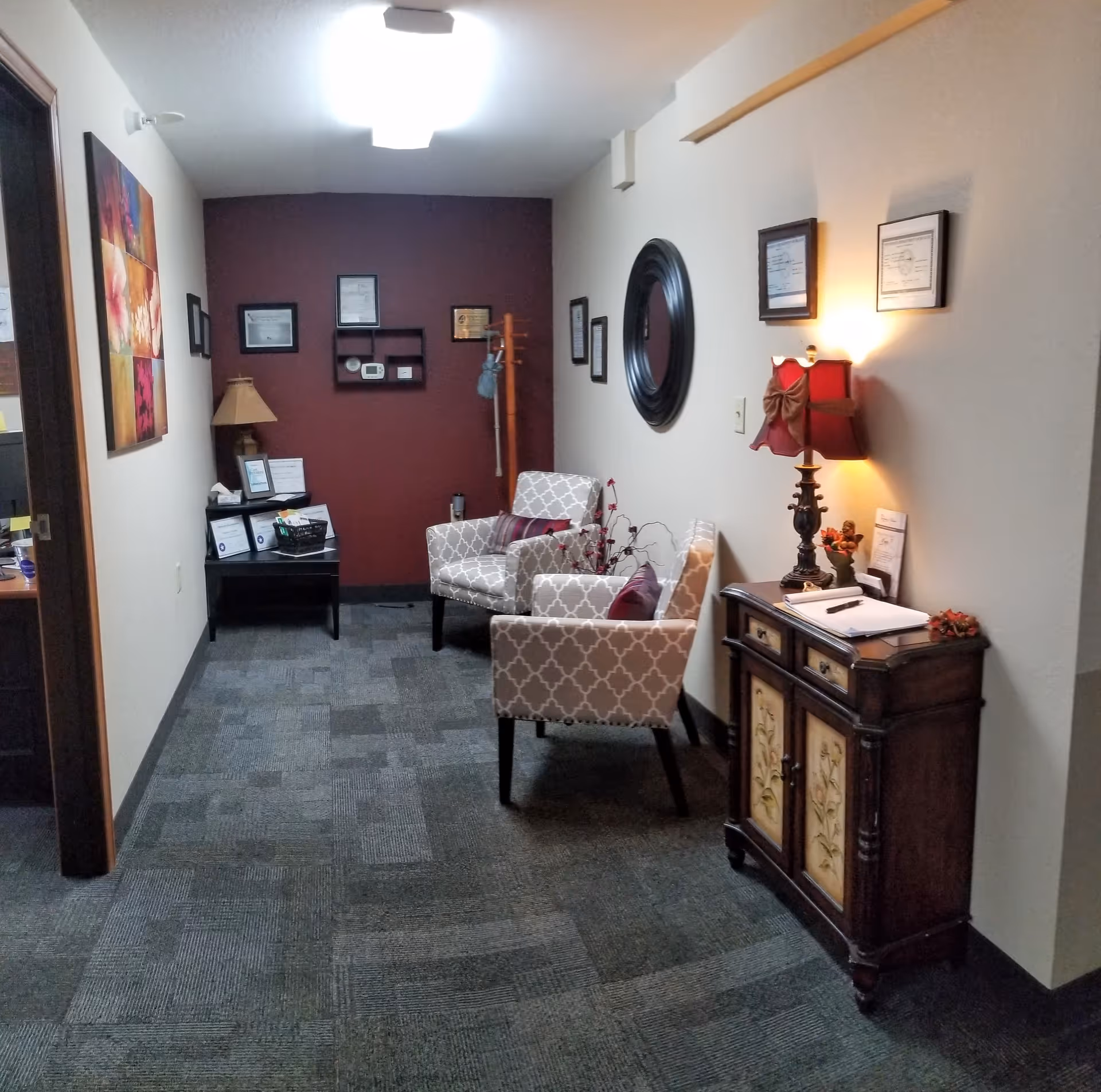 A small furnished waiting area with two patterned armchairs, side tables, lamps, wall art and a maroon accent wall.