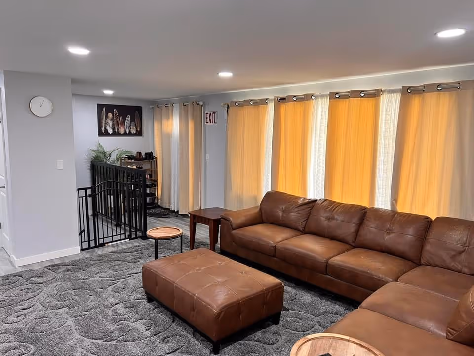 A cozy living room with a large brown leather sectional sofa and a matching ottoman on a gray patterned carpet. The room has several windows covered with beige curtains, and there are two small wooden side tables. A wall clock and a framed artwork featuring feathers are visible on the light gray walls. There is a black safety gate near a staircase and an exit sign above a door in the background.