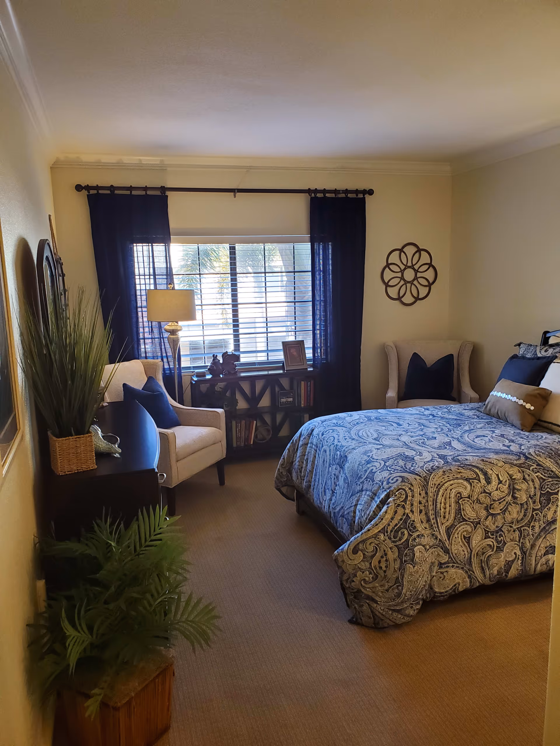 A cozy bedroom with a bed covered in a blue and beige paisley patterned comforter, two beige armchairs with dark blue pillows, a window with dark blue curtains, a small bookshelf with books and decorative items, a floor lamp, and green plants in the corner.