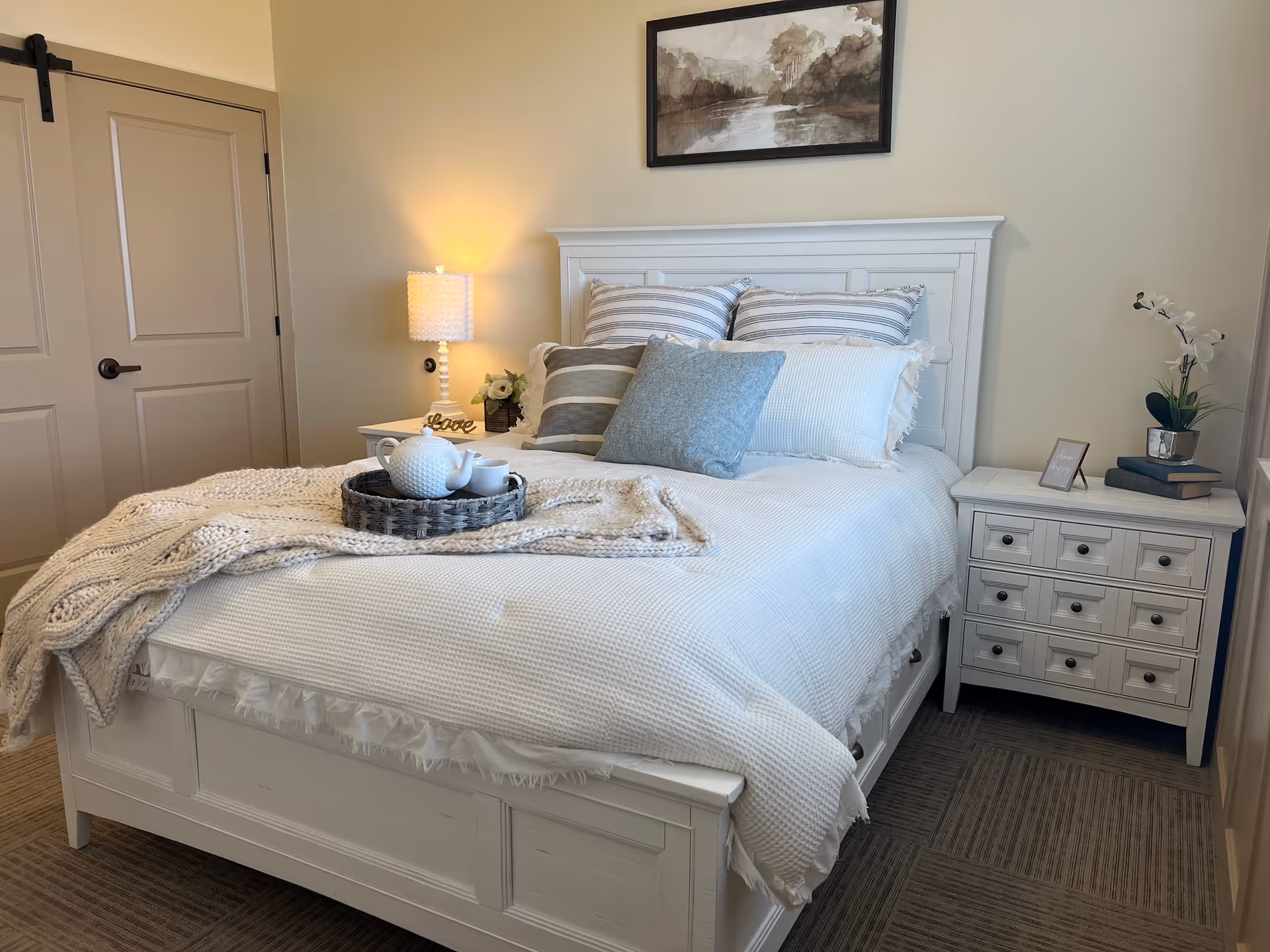 Cozy, well-lit bedroom featuring a white wooden bed with striped pillows, a knit throw and bedside tables with a lamp and decor.