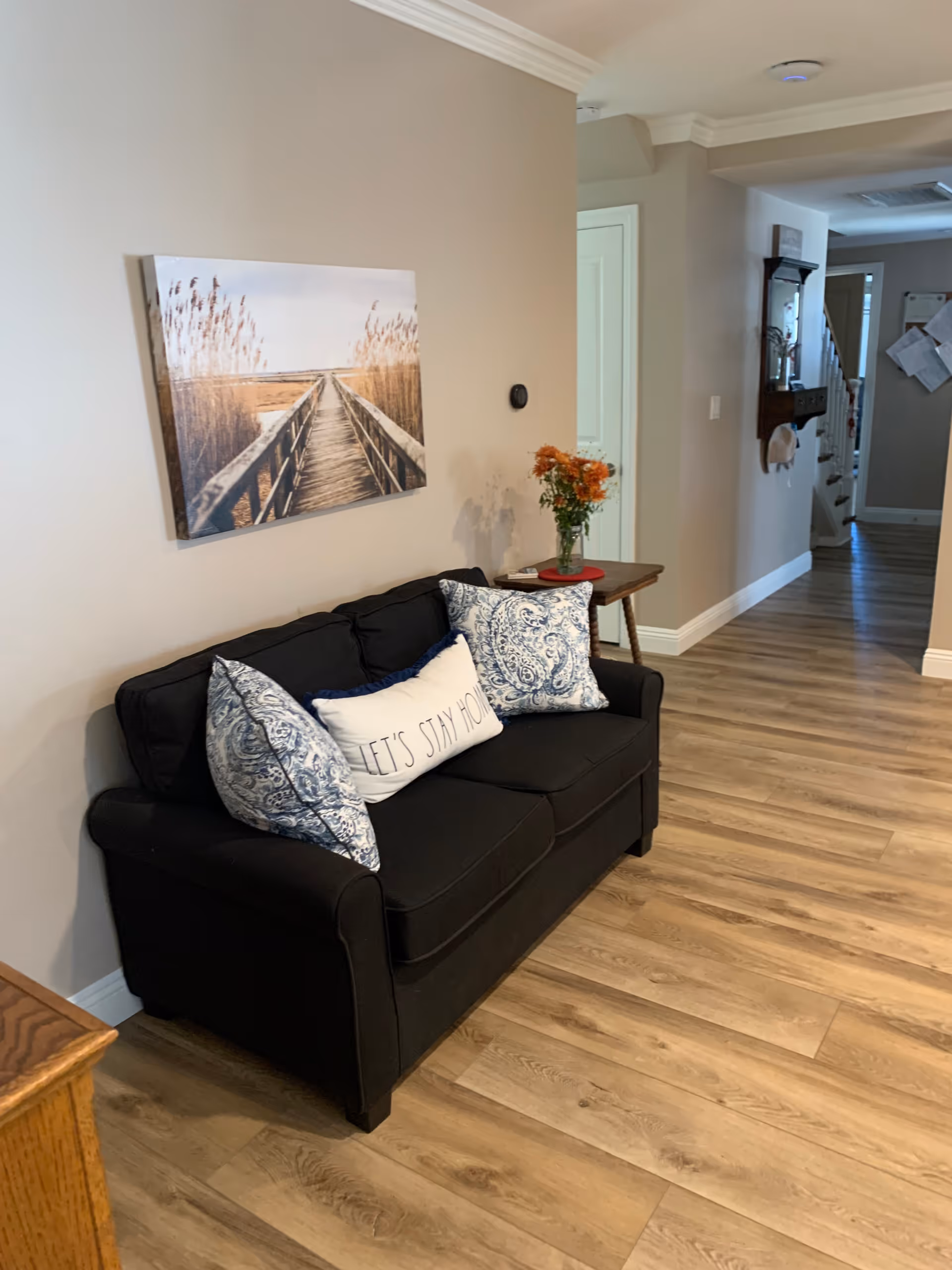A cozy interior hallway with a black couch adorned with three decorative pillows, one of which says 'LET'S STAY HOME'. Above the couch is a wall-mounted picture of a wooden boardwalk leading through tall grasses. A small wooden side table next to the couch holds a vase with orange flowers. The floor is light wood, and the hallway extends into the background with a staircase and some wall-mounted items visible.