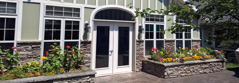 Exterior view of a building with light green siding and stone accents. There are two white-framed glass doors in the center, flanked by large windows. In front of the building, there are raised stone flower beds filled with colorful flowers and greenery. A tree branch partially shades the right side of the image.