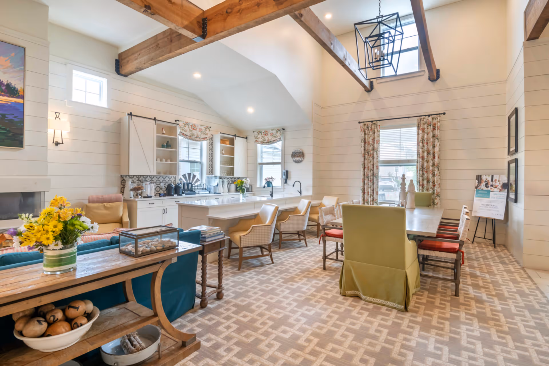 A bright and spacious interior room with high ceilings and exposed wooden beams. The room features a kitchen area with white cabinets, a large island with seating, and a dining table with various chairs. There are windows with floral curtains, a patterned carpet, and decorative items including flowers and framed pictures on the walls.