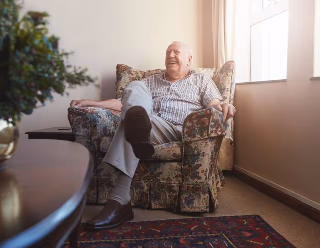 An elderly man sitting comfortably in a floral-patterned armchair in a sunlit room, smiling and looking relaxed. The room has a window with light curtains, a patterned rug on the floor, and a wooden table with a plant in the foreground.