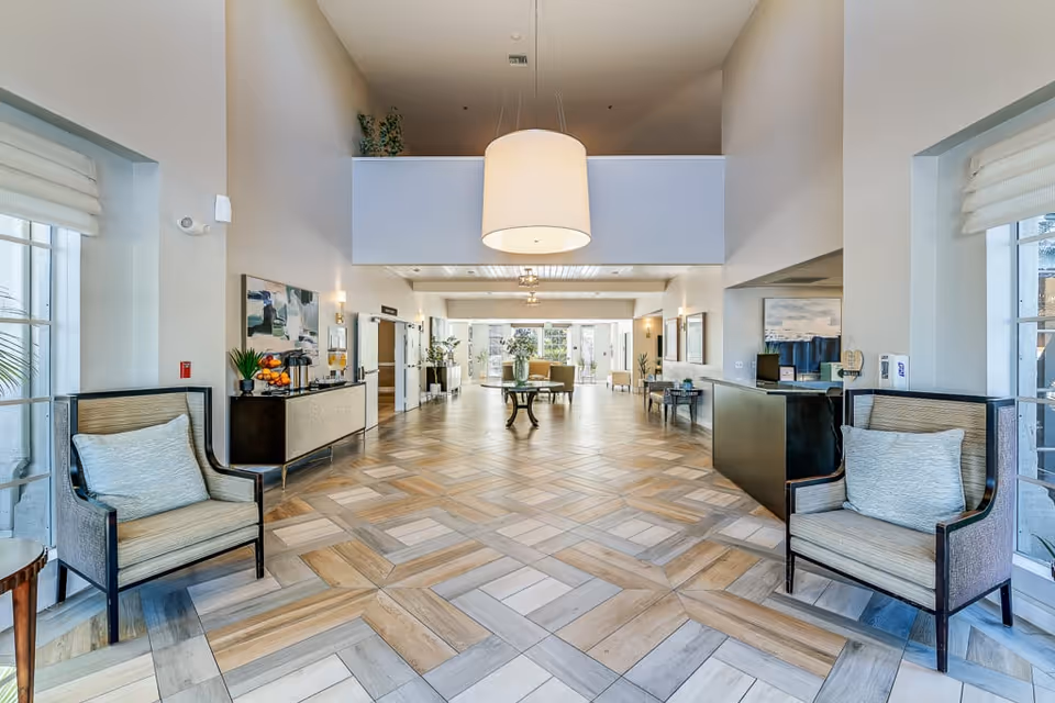 Spacious and well-lit senior living facility lobby with high ceilings, patterned tile flooring, two armchairs with cushions near large windows, a central round table with a vase of flowers, and a reception desk on the right side.