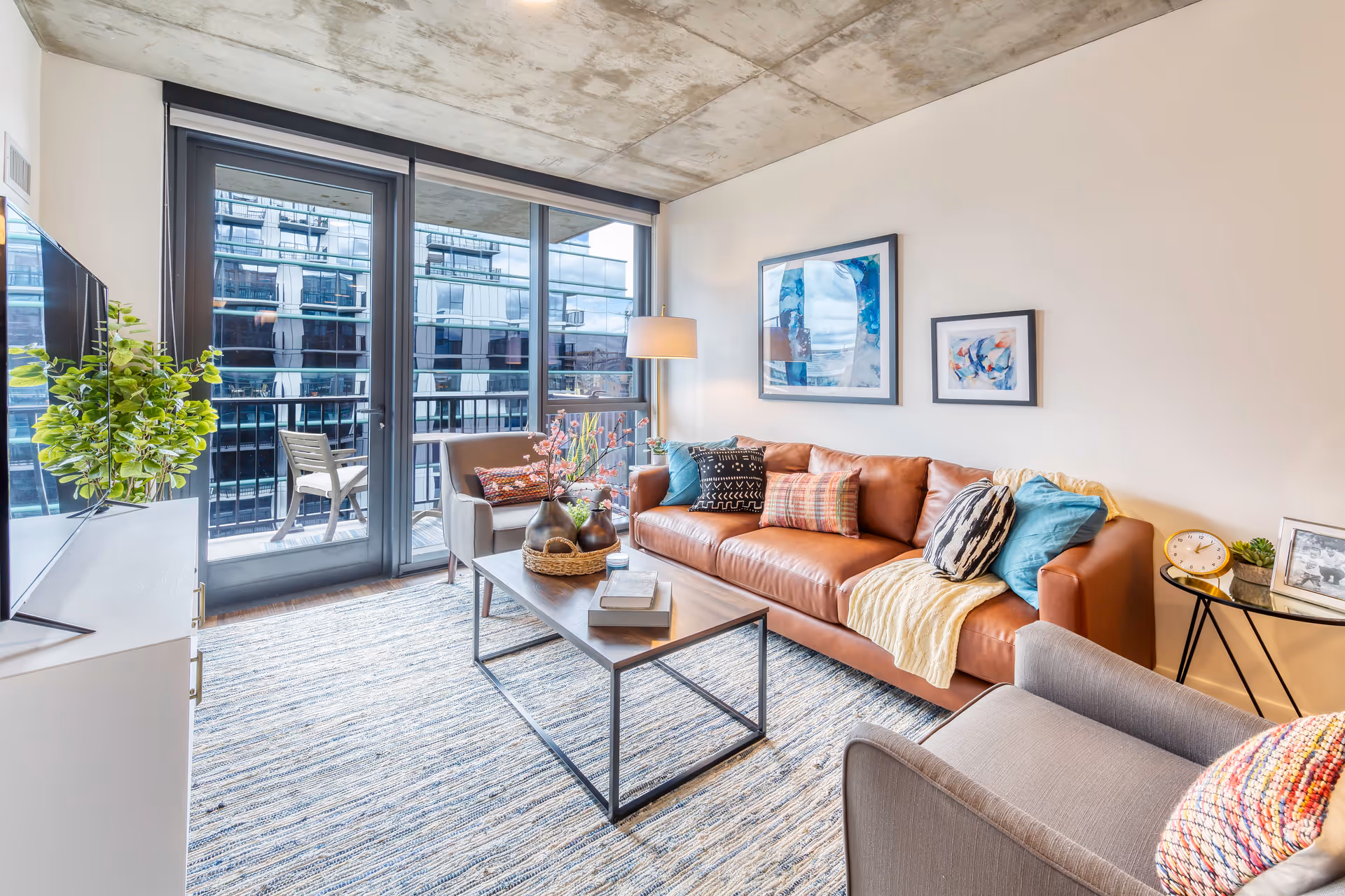 Modern living room with a brown leather sofa adorned with colorful pillows and a yellow throw blanket, a gray armchair with a multicolored pillow, a wooden coffee table with decorative vases and books, a floor lamp, and large floor-to-ceiling windows leading to a balcony with a chair. The room has a textured rug and two abstract paintings on the wall.