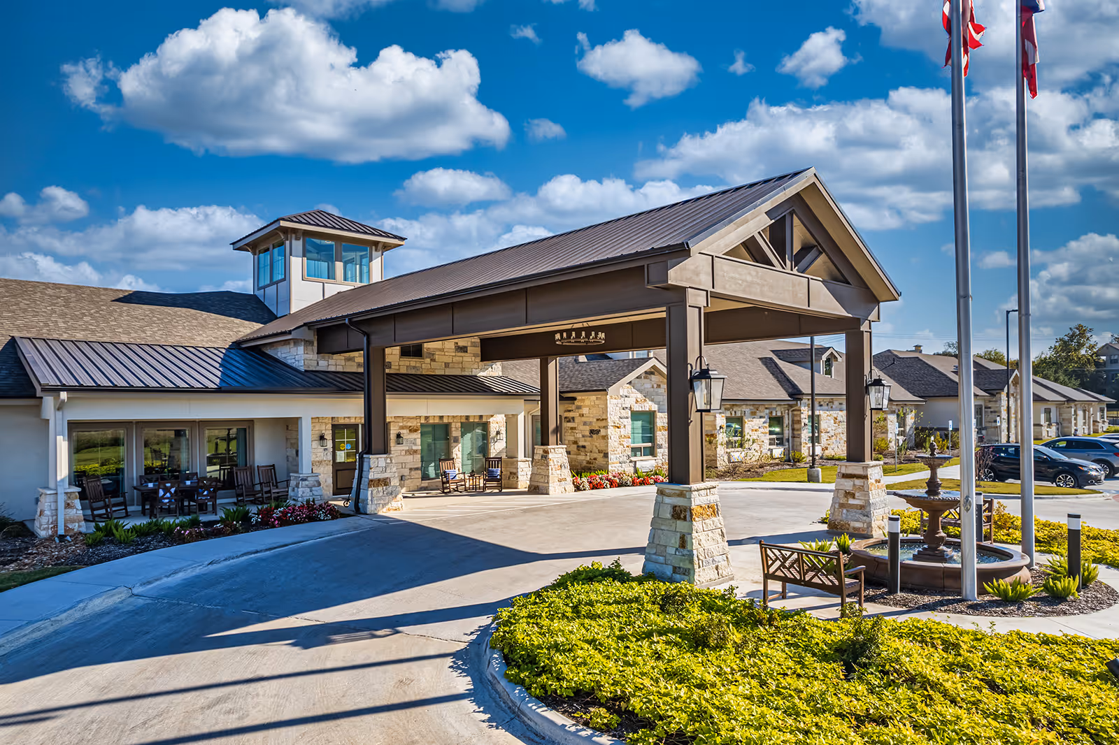 Front entrance of a senior living facility with a covered porte-cochere, flagpoles, fountain, benches, and landscaped grounds under a blue sky.