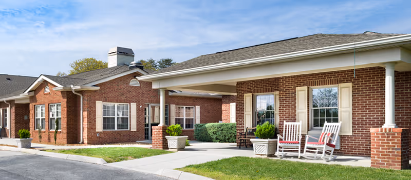 Exterior view of a single-story brick assisted living facility with white trim and columns. The building has multiple windows with beige shutters and a covered porch area with white rocking chairs and potted plants. The sky is clear with a few clouds.