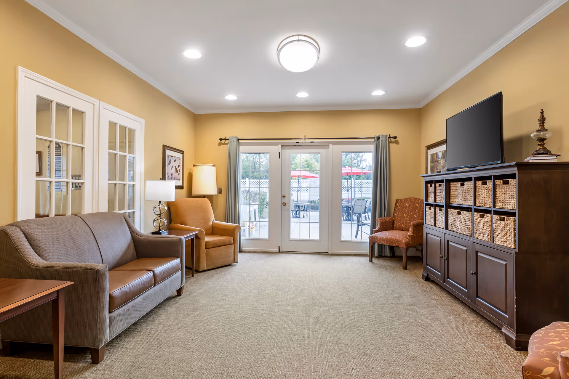 A cozy living room with beige walls and carpet, featuring a gray sofa, two armchairs, a wooden cabinet with woven baskets, a flat-screen TV, and a lamp. French doors with glass panes lead to an outdoor patio with tables and red umbrellas.