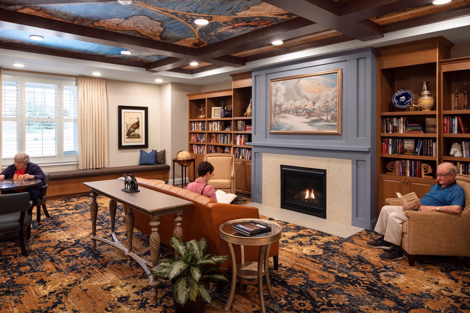A cozy living room in a senior living facility with three elderly people engaged in quiet activities. One person is sitting at a table near a window with beige curtains, another is seated on a brown leather sofa reading a book, and a third person is sitting in an armchair reading a newspaper. The room features built-in wooden bookshelves filled with books and decorative items, a fireplace with a framed painting above it, and a patterned carpet. The ceiling has decorative beams and a mural.