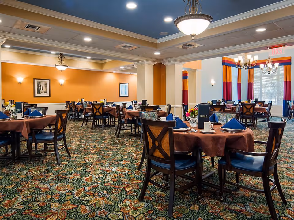 A spacious dining room with multiple round tables covered with brown tablecloths and set with blue napkins, cups, and menus. The room features patterned carpet, warm orange and white walls, framed artwork, and ceiling lights including chandeliers. Large windows with colorful curtains allow natural light into the room.