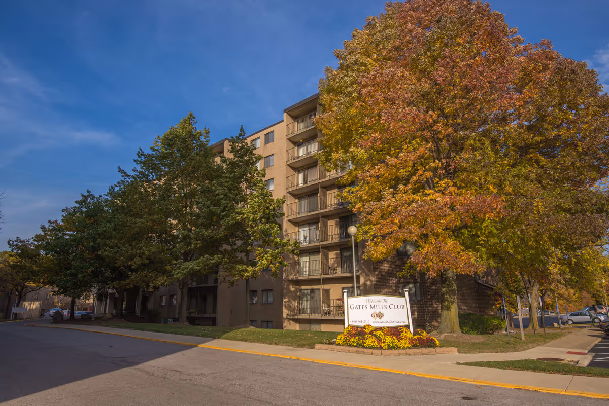 Exterior view of a multi-story senior living facility building named Gates Mills Club, surrounded by trees with autumn foliage and a clear blue sky. A sign in front of the building reads 'Welcome to Gates Mills Club' with contact information.