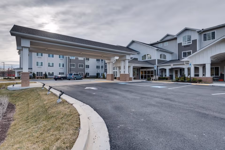 Front exterior of a multi-story senior living building showing a covered porte-cochere, entrance, and parking area.