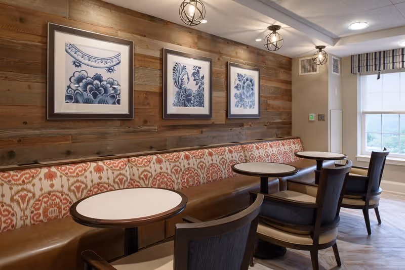 A cozy dining area with a long cushioned bench featuring a patterned fabric backrest and leather seat, accompanied by round tables and wooden chairs. The wall behind the bench is covered with wooden panels and decorated with three framed blue and white floral artworks. Two modern ceiling light fixtures hang above, and a window with a striped valance lets in natural light.
