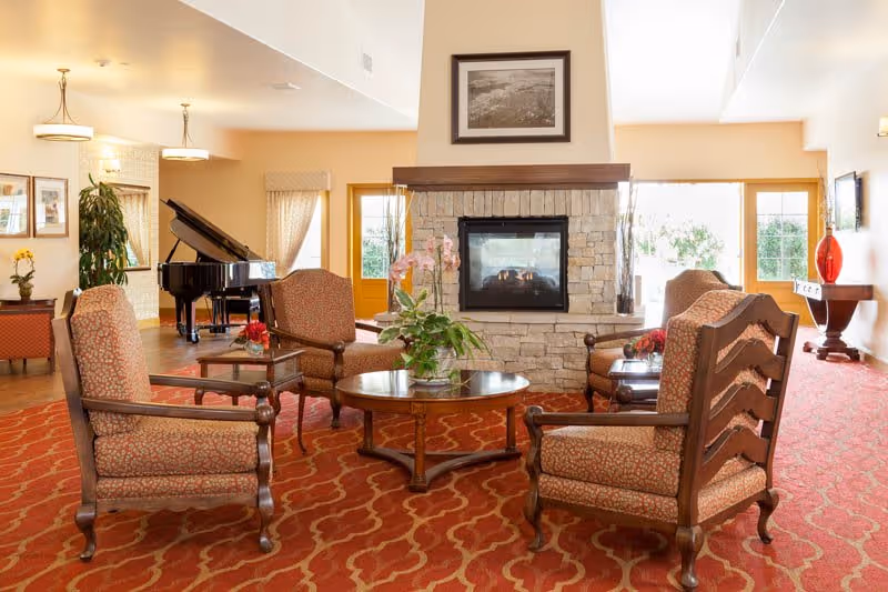 A cozy living room area with four upholstered armchairs arranged around a round wooden coffee table with a plant centerpiece. A stone fireplace with a framed picture above it is in the center background. To the left, there is a black grand piano and some framed pictures on the wall. The room has large windows and doors letting in natural light, and the floor is covered with a red patterned carpet.