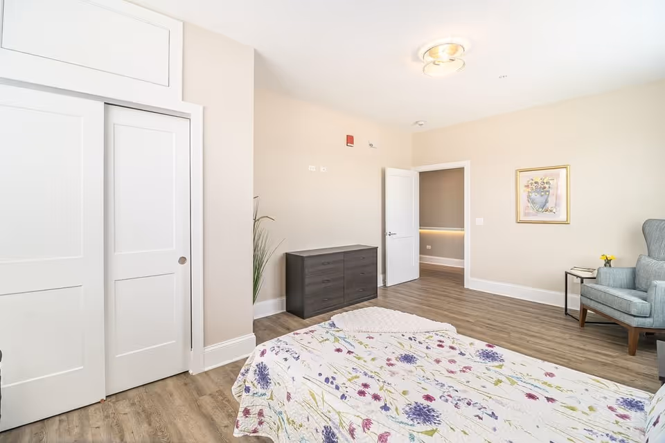 A bright and spacious bedroom with light beige walls and wood flooring. The room features a bed with a floral patterned bedspread, a dark wooden dresser, a light blue upholstered armchair next to a small side table with a yellow flower vase, and a framed floral painting on the wall. There is a white door open to an adjacent room and a white closet with sliding doors.