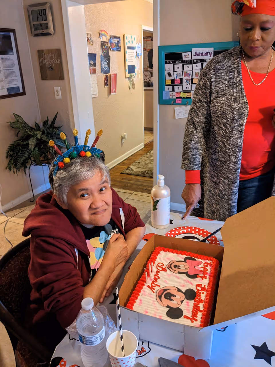 An older woman wearing a colorful birthday headband sits at a table smiling beside a Mickey Mouse birthday cake while another woman stands nearby in a communal dining area.