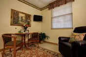 A cozy room with a small round wooden table and two wooden chairs with cushions. A flat-screen TV is mounted on the wall above a potted plant. There is a large window with blinds and a decorative valance. A comfortable dark leather armchair with a patterned pillow is positioned near the window. The room has light-colored walls and a patterned area rug covering the floor.