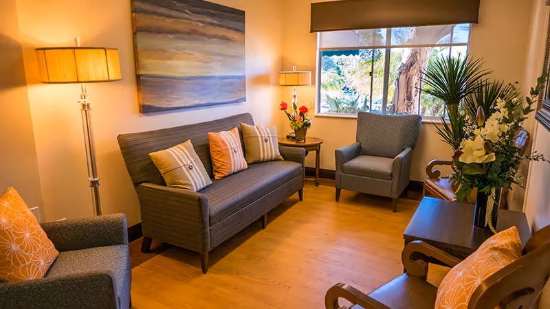 A cozy living room with a gray striped sofa adorned with four decorative pillows, two armchairs with orange floral cushions, a wooden side table with a vase of flowers, two standing lamps, a large window with a view of trees outside, and a painting on the wall above the sofa.