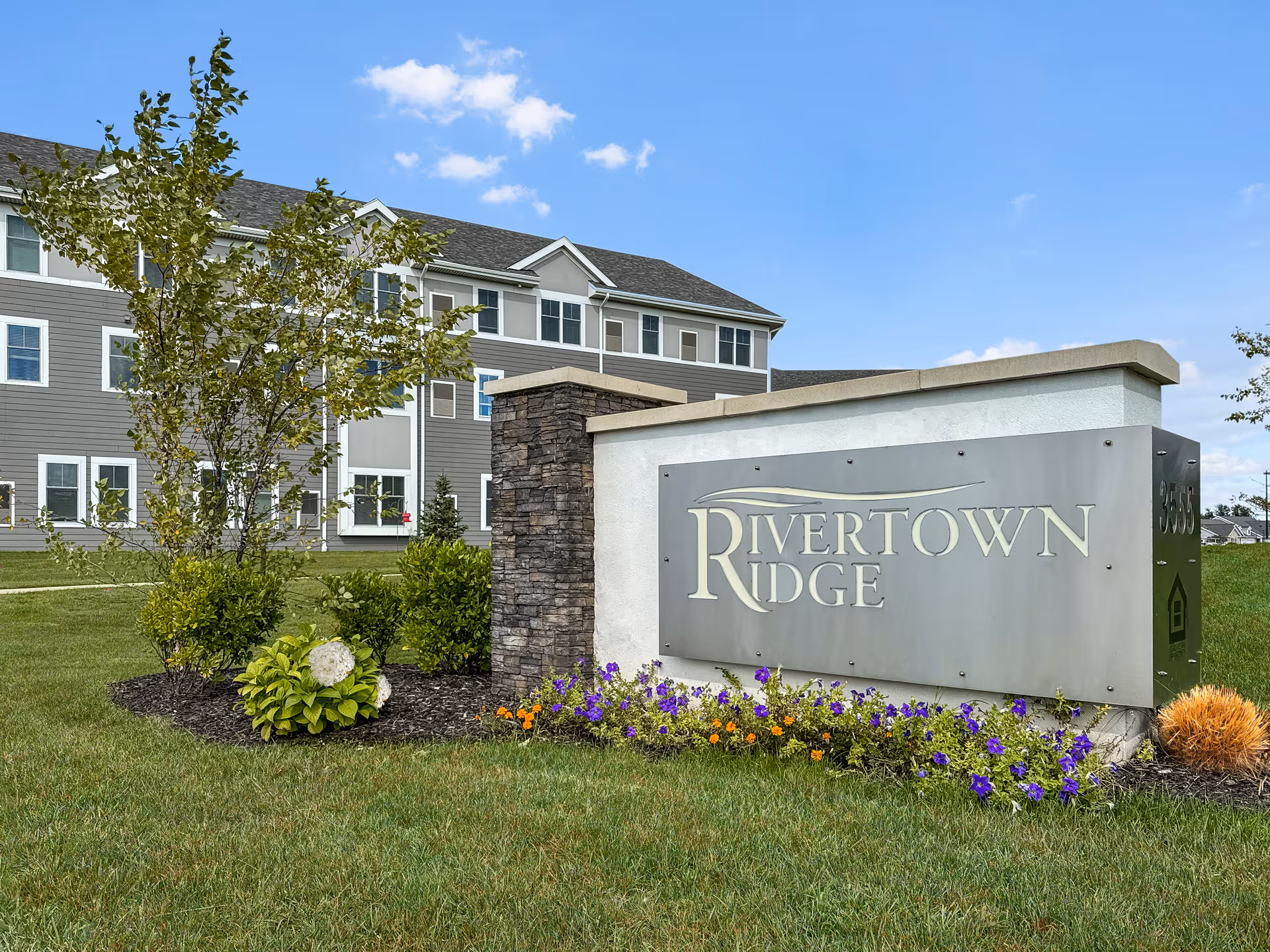 Outdoor view of Rivertown Ridge senior living facility sign with a landscaped garden featuring green grass, small bushes, and purple and orange flowers. The building with multiple windows is visible in the background under a blue sky with scattered clouds.