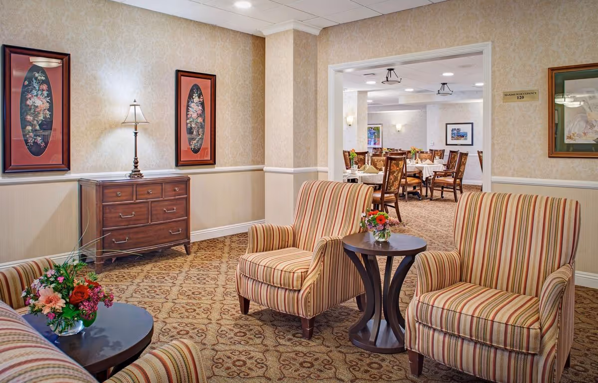 A cozy sitting area with two striped armchairs and a small round table with a flower vase. Behind the sitting area is a wooden chest of drawers with a lamp on top, flanked by two framed floral paintings. Through an open doorway, a dining room with tables and chairs is visible.